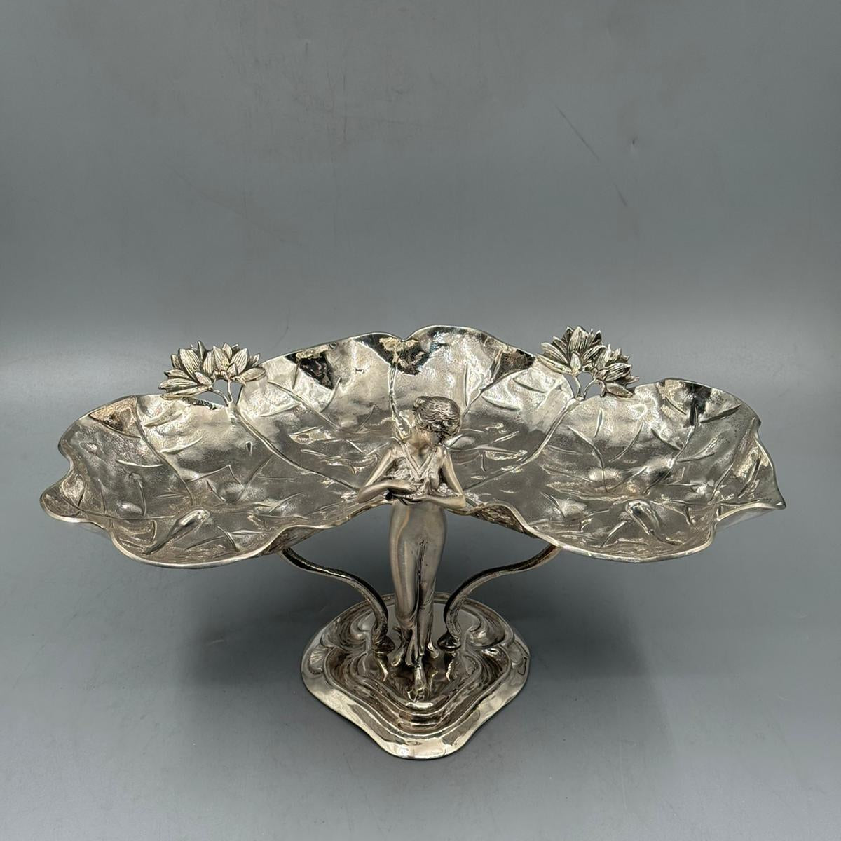 A vintage silver centerpiece depicting a female figure supporting two lotus leaf-shaped trays adorned with lotus flowers.