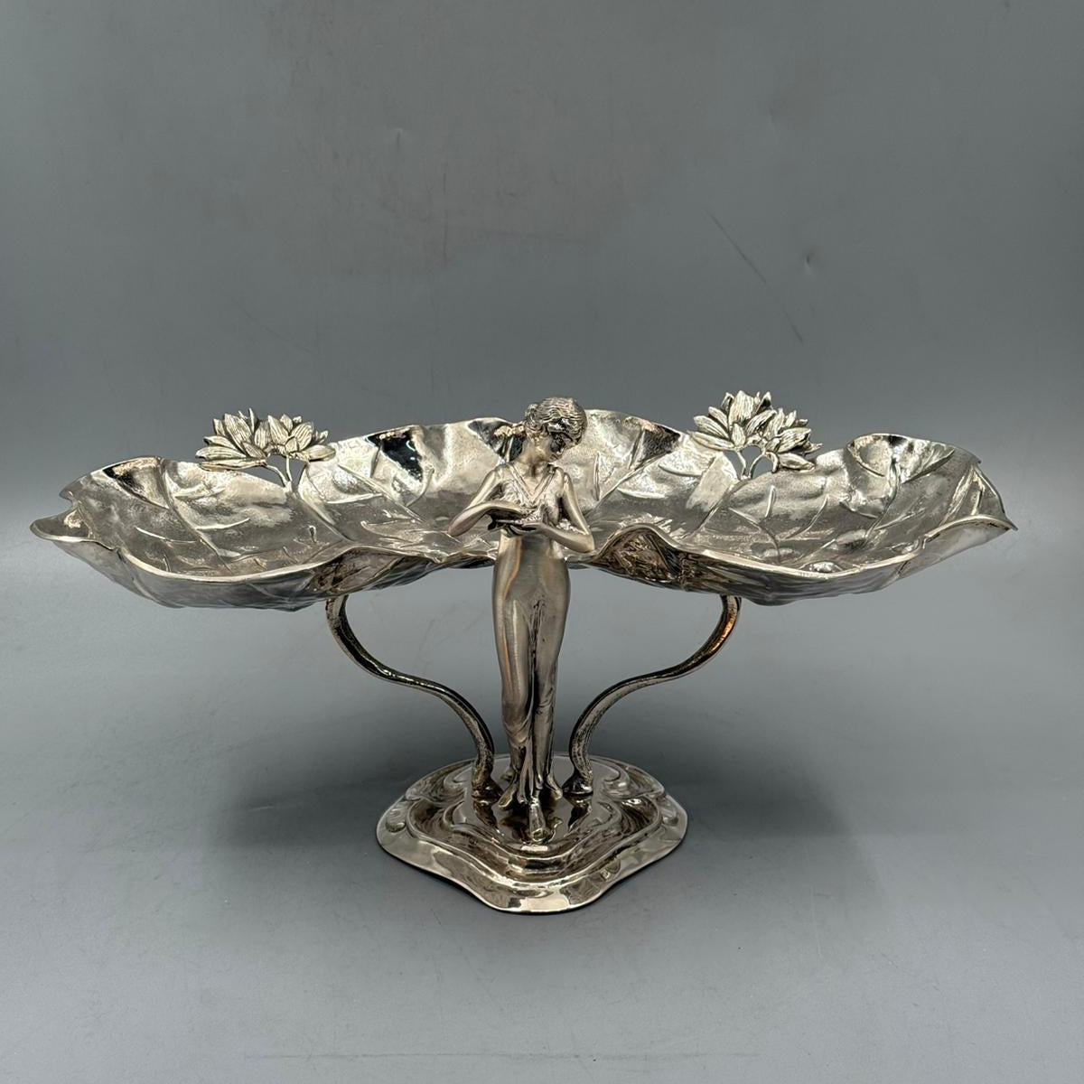 An ornate silver decorative centerpiece featuring a standing female figure holding two large lotus leaf trays with flower details.
