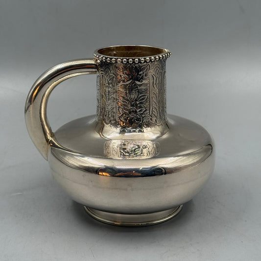 A vintage silver water jug featuring a wide rounded base, ornate floral engravings on the neck, and a curved handle.