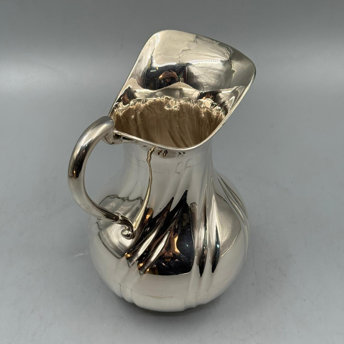 A vintage-style silver pitcher featuring a wide rounded body, decorative curves, and a polished mirror-like surface.