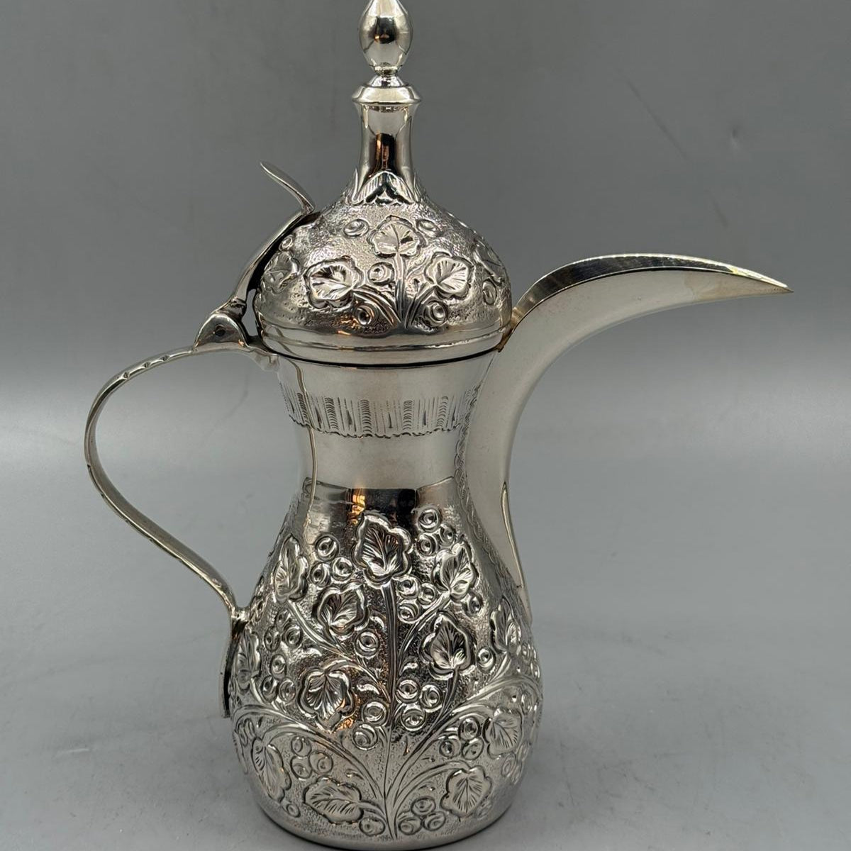 A shiny silver dallah pot with ornate flower motifs engraved on the body and lid.