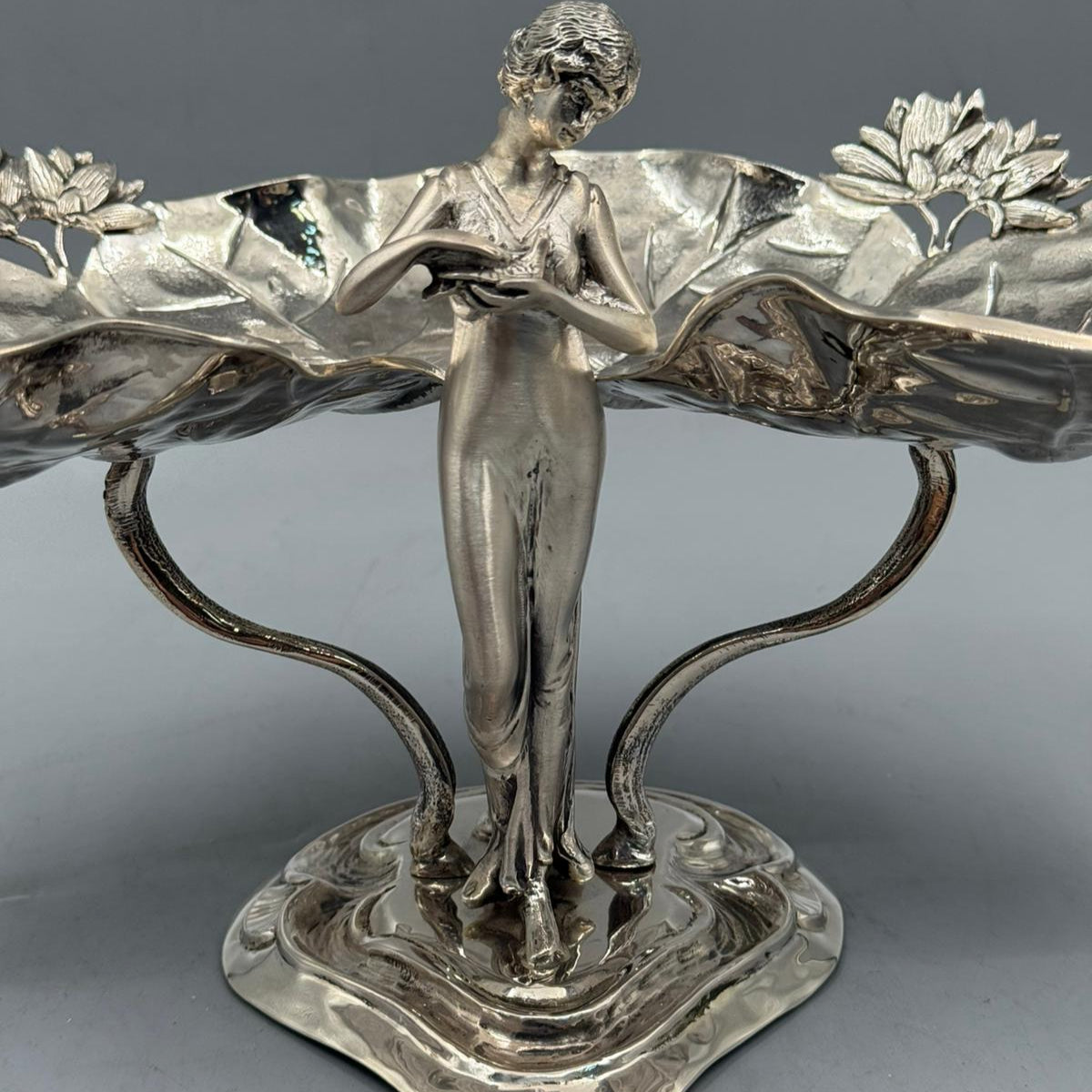 A polished silver centerpiece showing a standing woman figure holding up twin lotus leaf trays with detailed floral patterns.