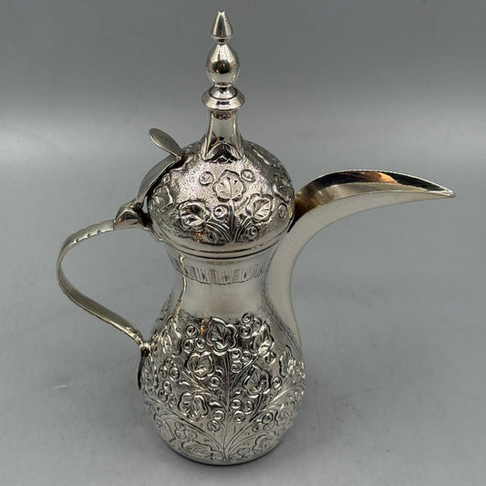 A traditional silver dallah coffee pot decorated with detailed floral patterns and a tall pointed lid.
