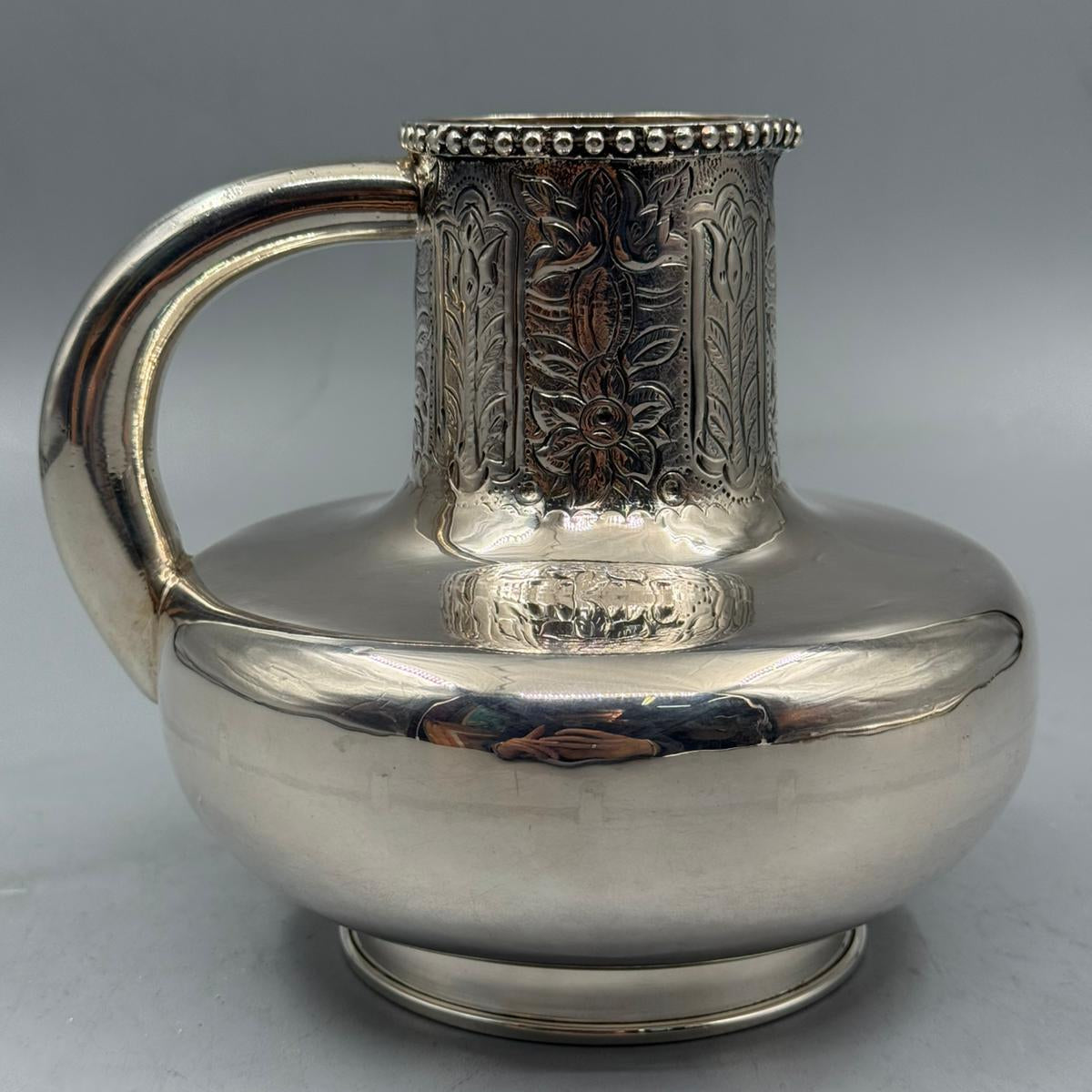 A shiny silver jug with a wide rounded shape, engraved floral motifs around the neck, and a smooth polished finish.