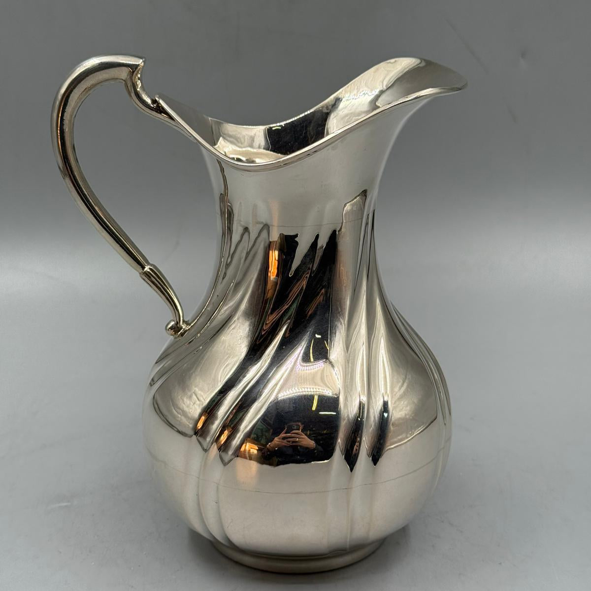 A shiny silver jug featuring a bulbous base, curved handle, and a wide square-shaped spout.