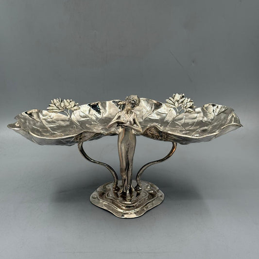 An ornate silver decorative centerpiece featuring a standing female figure holding two large lotus leaf trays with flower details.