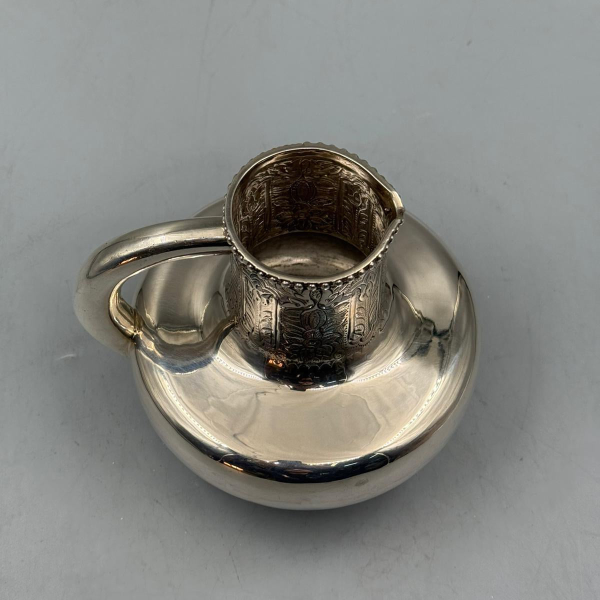 An antique-style silver pitcher with intricate floral engravings on the upper neck and a wide circular body.