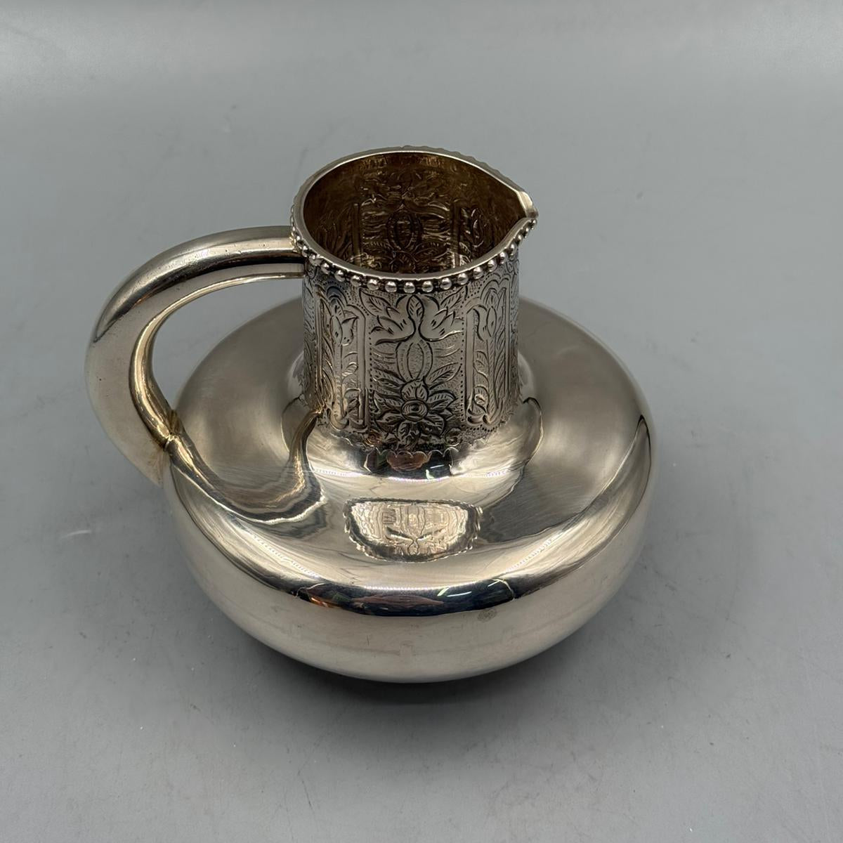 A polished silver jug with a flat rounded body, decorated neck engravings, and a sturdy curved handle.