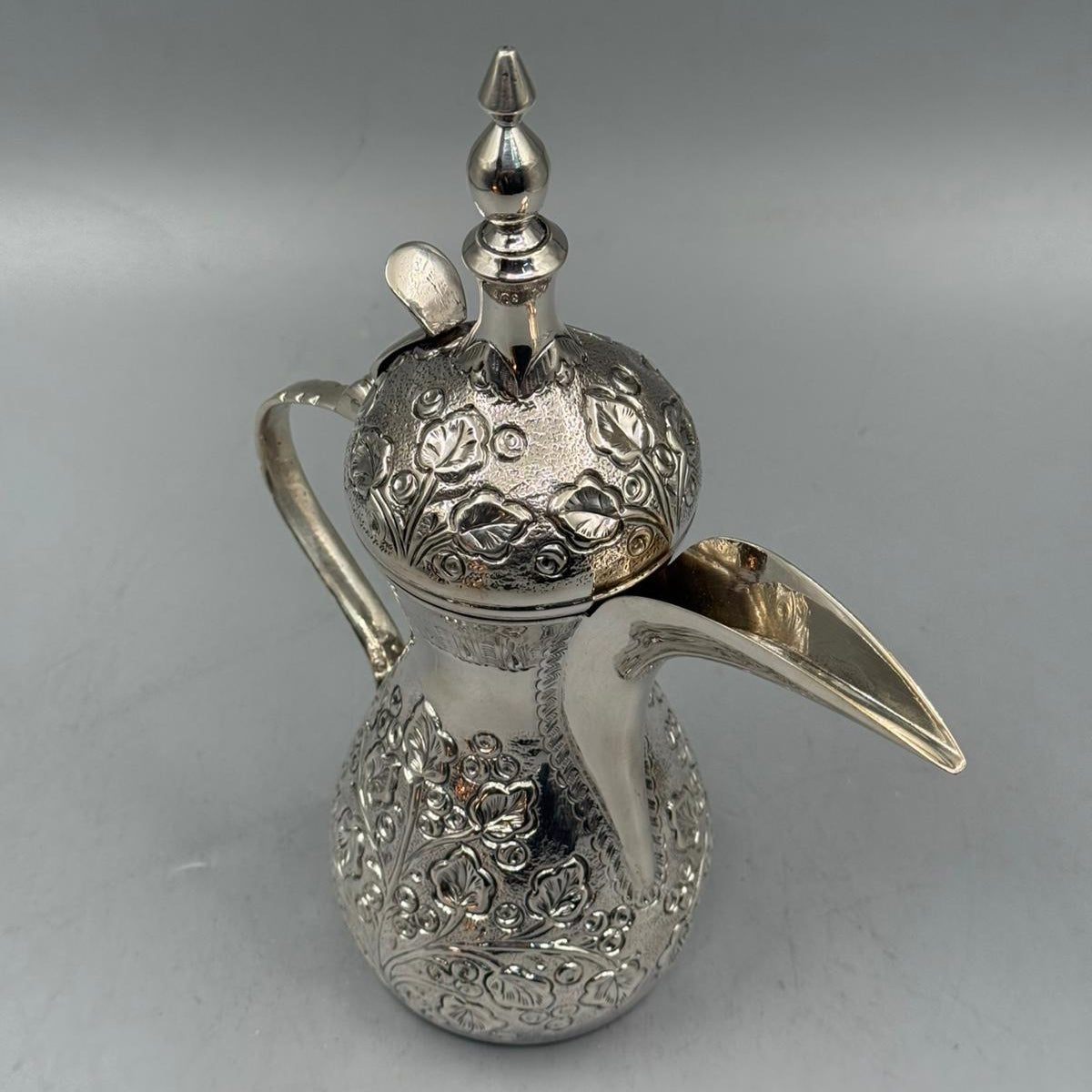 An antique silver Arabic coffee pot featuring floral carvings, a curved handle, and a long spout.