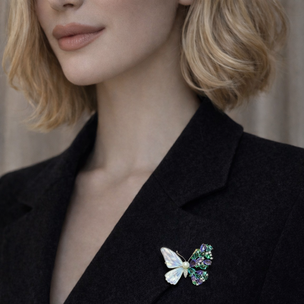 A stunning asymmetrical butterfly brooch in a black velvet box. One side features iridescent mother of pearl wings, while the other is encrusted with green and purple crystals, joined by a delicate pearl center.