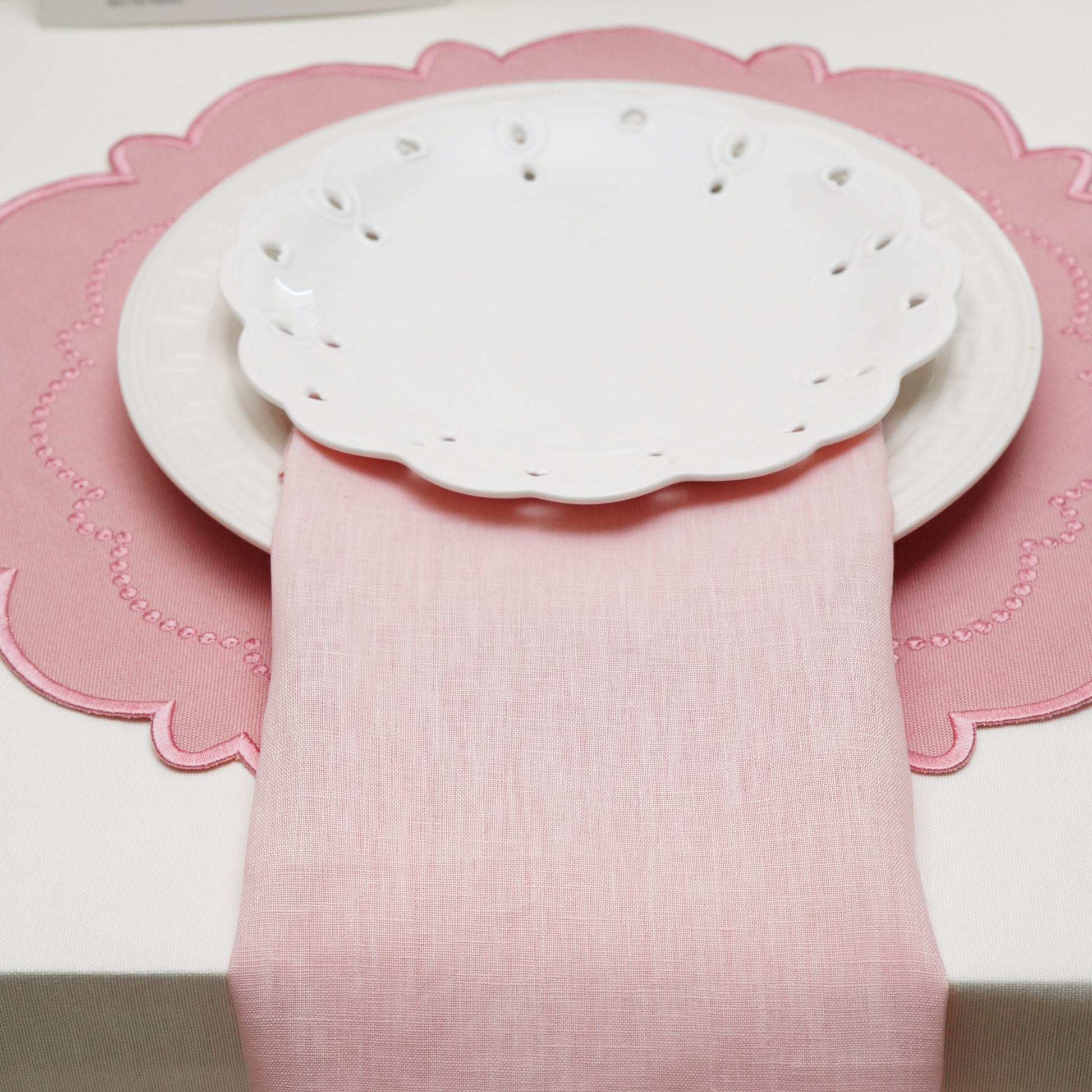 A beautiful blush pink linen hemstitch cloth napkin elegantly draped vertically under stacked white scalloped plates on a matching pink placemat.