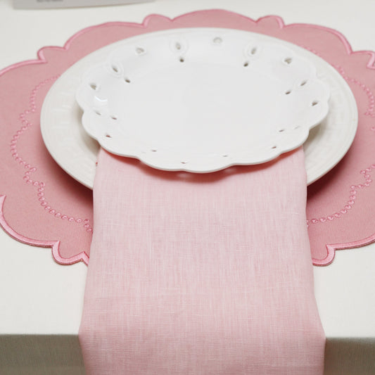 A beautiful blush pink linen hemstitch cloth napkin elegantly draped vertically under stacked white scalloped plates on a matching pink placemat.