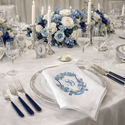 Grand wedding table setting featuring a white cloth napkin with a custom blue floral crest monogram, placed on a silver-rimmed plate with navy flatware and elegant blue and white floral centerpieces.