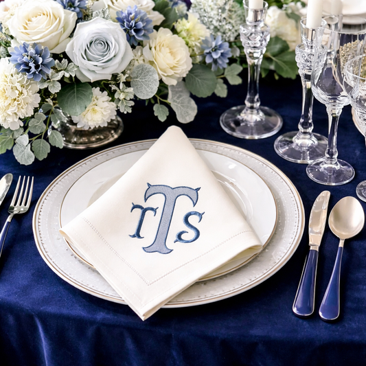 Elegant navy and silver table setting featuring a white cloth napkin with a custom classic blue three-letter monogram, resting on a layered plate over a rich navy velvet tablecloth alongside crystal glassware.