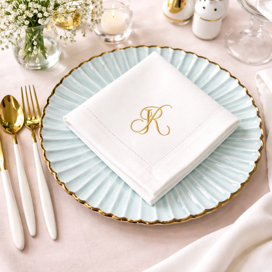 Elegant table setting featuring a folded white cloth napkin with a custom gold monogram letter R, resting on a light blue scalloped plate with a gold rim, accompanied by white and gold flatware on a soft pink tablecloth.