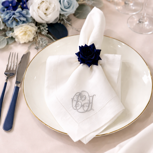 Elegant table setting featuring a folded white cloth napkin with a custom silver monogram, accented by a navy blue rose napkin ring on a gold-rimmed plate alongside navy flatware.