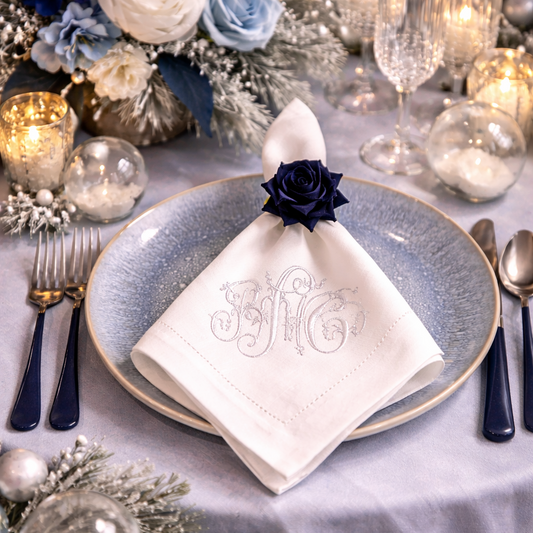 Elegant winter table setting featuring a white cloth napkin with a custom silver monogram, adorned with a navy blue rose napkin ring on a light blue textured plate.