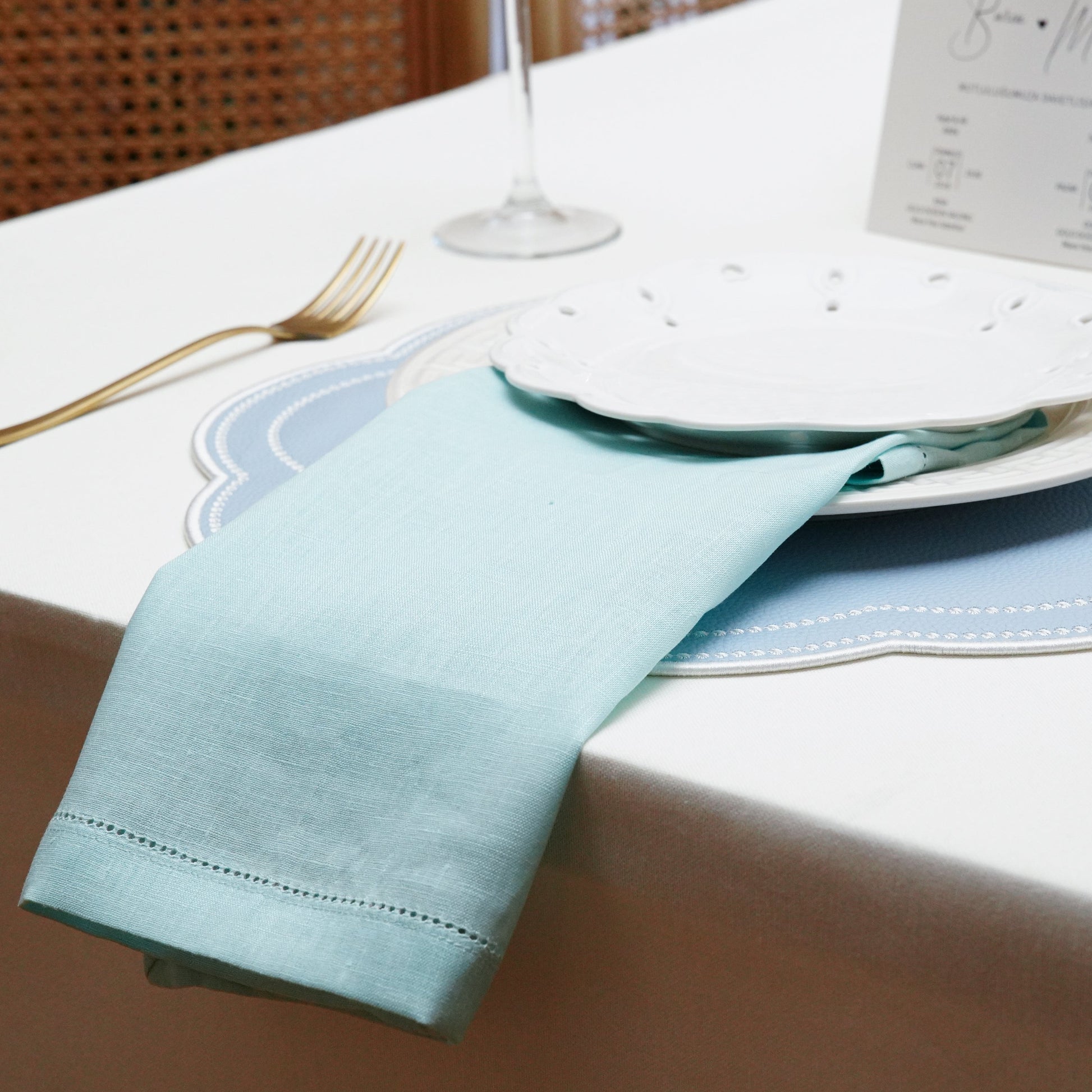 A romantic spring wedding table decor showcasing light blue table linens, a delicate hemstitched napkin, white floral centerpieces, and a personalized wedding invitation.