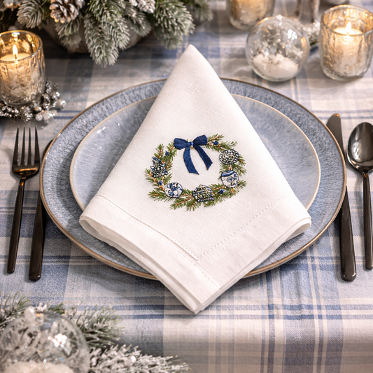 Elegant winter holiday table setting featuring a white cloth napkin with a custom embroidered pine wreath, blue ornaments, and a navy bow, resting on a light blue plate over a plaid tablecloth.