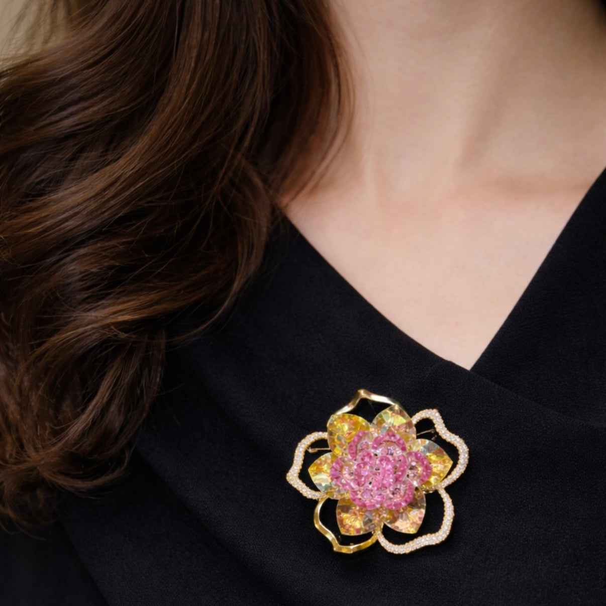 A woman wearing an elegant black dress adorned with a statement gold-plated flower brooch. The floral pin features vibrant yellow and pink stones with pave-set clear crystals on the outer gold wireframe petals.