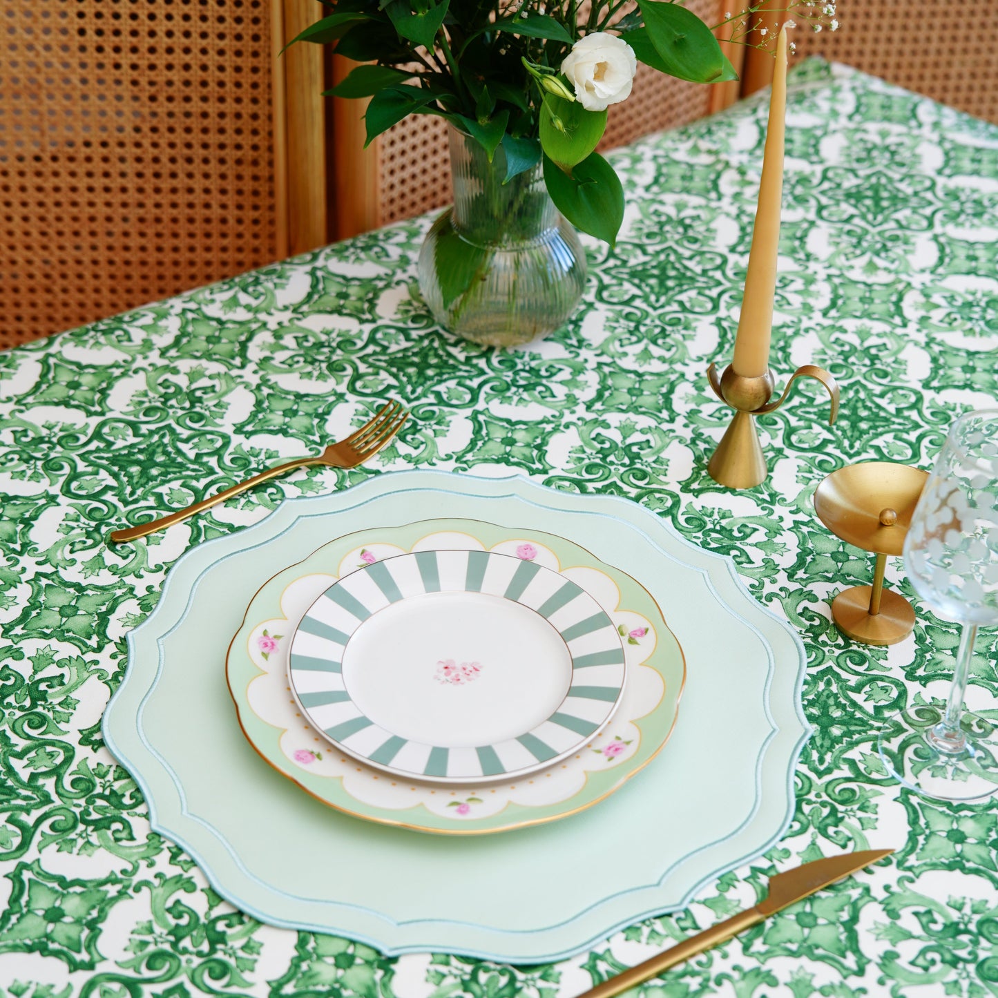 Green and white damask tablecloth styled with plates, gold flatware, candleholder, and a flower vase for an elegant tablescape