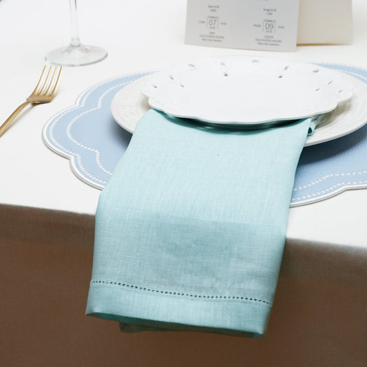 A light blue linen hemstitch cloth napkin elegantly draped under a scalloped white plate, set on a matching blue scalloped placemat for a wedding reception.