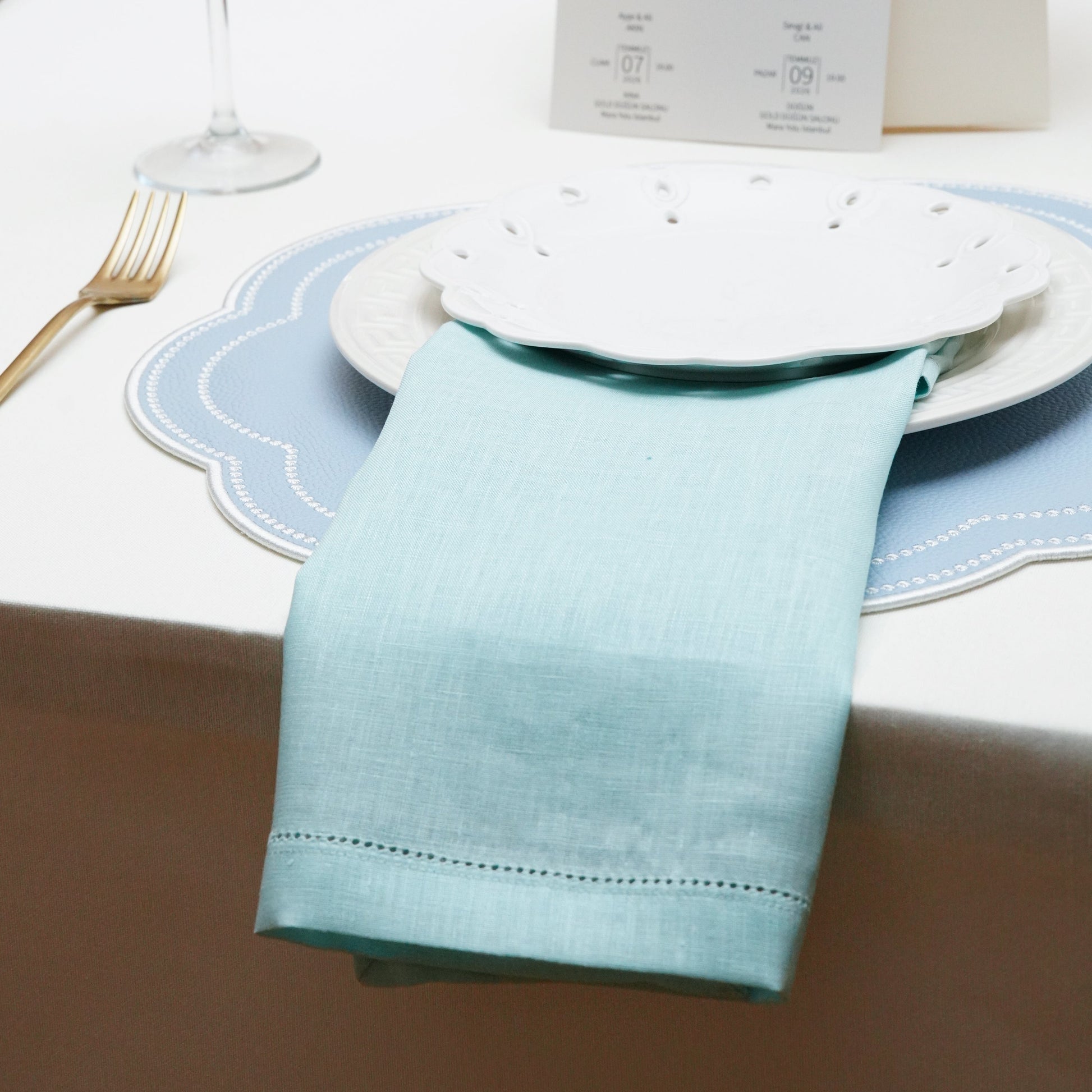 Elegant table setting featuring a pastel blue linen napkin and a scalloped placemat with white stitching, accompanied by a gold fork and polka dot glass.