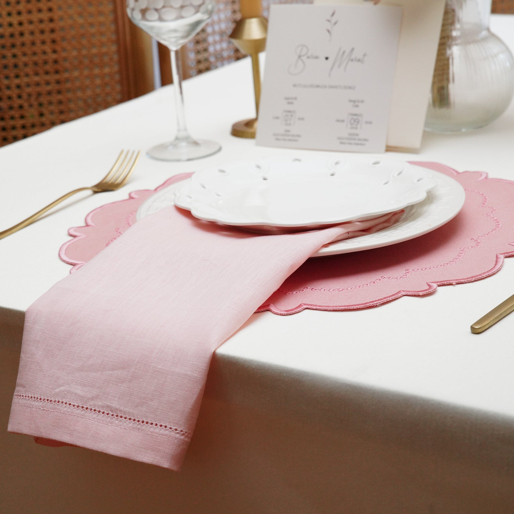 A matching set of a pink scalloped embroidered placemat and a light pink linen napkin, arranged beautifully with fine white dinnerware.